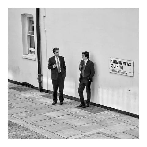This is a black and white square format image of two men in suits taking a cigarette break on the corner of a London street. Their dark suits sit in stark contrast to the pale render of the building they stand next to. They are centred in the frame, the man to the right leaning on the wall which sits to an angle from the viewer, and the man to the left turning to him in conversation. Taken in London with a Sony RX100.