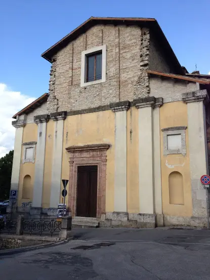 The image features a building that appears to be a church or a historical structure. It has a façade with a mix of stone and plaster, displaying a yellowish color. The front entrance is framed by a wooden door, and there are a few architectural details, such as columns. The setting seems to be an urban area, with some street elements visible. The sky is partly cloudy, suggesting a clear day.

Image Credits: Wikimedia / Manuelarosi / CC BY-SA 3.0