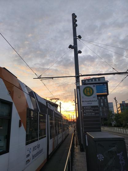Links im Bild ist eine Seite einer Straßenbahn zu sehen, rechts ein leeres Bahngleis. Darüber der Himmel in gelb, orange, rot und blau.