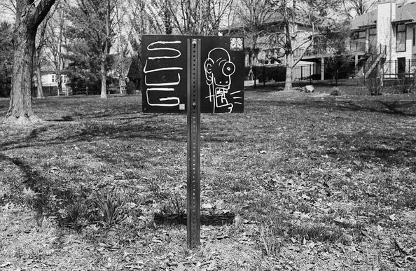 A sign posted in the park--someone  has painted the back of it black and scrawled graffiti in white in a sort of scratchboard technique.