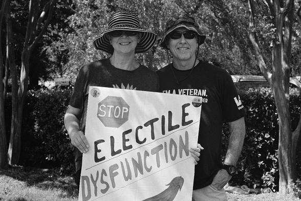 A couple, the husband is a veteran according to his shirt, and a sign that says “stop electile dysfunction”