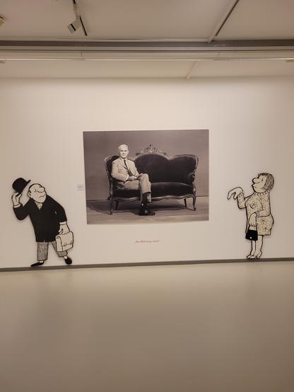 Auf dem Foto sieht man links und rechts jeweils eine Zeichenfigur von Loriot.
In der Mitte hängt ein Foto auf dem die berühmte Couch  und Loriot selbst zu sehen ist.