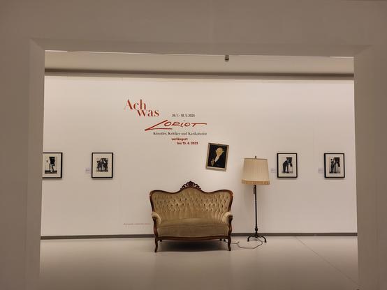 Zu sehen ist eine Nachbildung der berühmten Couch von Loriot, daneben steht eine Stehlampe.
Im Hintergrund sieht man mehrere Bilder die Loriot wiedergeben.