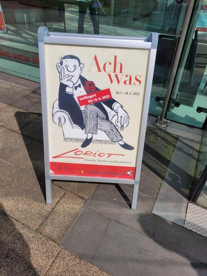 Vor dem Eingang zur Galerie steht ein Schild auf dem eine der Figuren von Loriot auf einem Sessel sitzt.
Darüber steht der Name der Ausstellung, "Ach was".