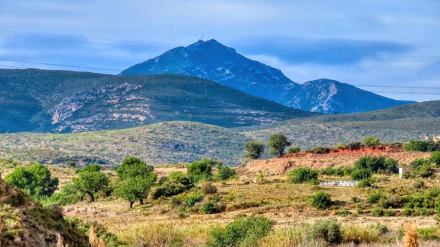 Aquesta imatge mostra un paisatge serè i muntanyós. En primer pla, hi ha vegetació baixa i dispersa, amb alguns arbres escampats pel terreny àrid. El color predominant és el marró daurat de la terra seca, en contrast amb el verd dels arbres i arbustos. Al centre i al fons, imponents muntanyes s'aixequen majestuosament, els seus vessants coberts de vegetació de color verd fosc que es torna blavós a les parts més altes. Una de les muntanyes destaca per la presència d'una petita estructura al seu cim, possiblement un transmissor o antena. El cel és d'un blau clar i una mica ennuvolat, aportant una sensació de calma i tranquil·litat a l'escena.