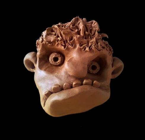 The image shows a sculpture made of plasticine of a cartoonish face with exaggerated features against a black background. It has large round hollow eyes, prominent ears, a textured wavy top resembling hair, a pronounced underbite with teeth showing, and a smooth rounded chin. The sculpture has a light brown/terracotta color and appears to be crafted in a whimsical, slightly grotesque style.
