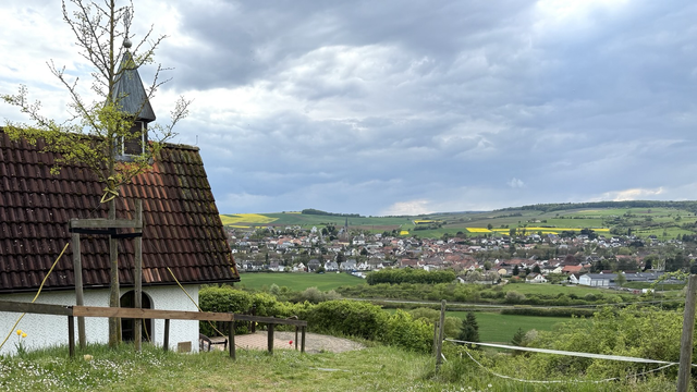 Eine Kapelle mit weißen Mauern und einem großen Ziegeldach sowie einem kleinen Türmchen im Vordergrund, in der Ferne ein Dorf und teils grüne, teils gelbe Felder. Am Himmel eine Wolkendecke.