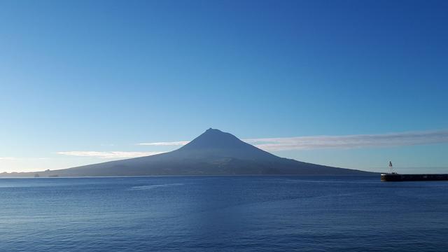 Ein klarer, sonniger Blick über das blaue Meer auf die Azoreninsel Pico mit ihrem markanten Vulkankegel, dem höchsten Berg Portugals. Der Vulkan ragt symmetrisch und majestätisch in den Himmel. Am rechten Bildrand ist ein kleiner Hafen mit einem rot-weißen Leuchtturm zu sehen. Der Himmel ist wolkenarm und strahlend blau, mit einem schmalen Wolkenband knapp über dem Horizont.