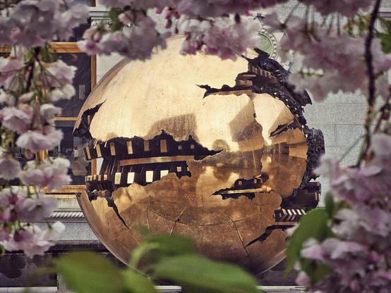 A partially obscured view of Arnaldo Pomodoro's "Sphere Within Sphere," a large, cracked golden globe reflecting surrounding buildings, framed by soft-focus pink cherry blossoms.