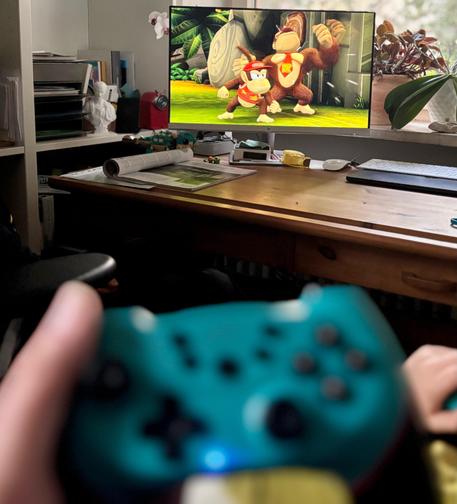Ein unscharfer Blick über zwei Hände mit einem türkisfarbenen Nintendo Switch Pro Controller im Vordergrund. Im Hintergrund sieht man einen Schreibtisch mit einem Monitor, auf dem Donkey Kong und Diddy Kong aus dem Spiel „Donkey Kong Country Returns HD“ in Siegerpose zu sehen sind.