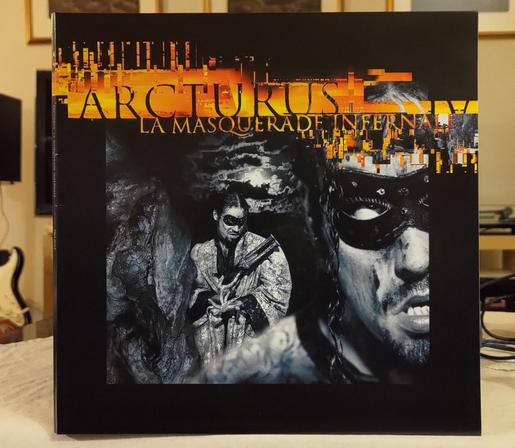ARCTURUS - La Masquerade Infernale LP cover. Band name and title in orange/orange background  on top, partly over  a black and white photo of two men (or maybe it two photos of the same person) in with masks (covering their eyes). One is a close-up of part of the face, and the other from a distance (holding an old gun)