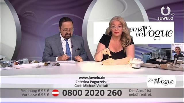 Description: The image shows a man and woman sitting at a table in front of a TV screen. The woman is wearing a black dress and the man is wearing spectacles. On the table there are papers, a cup, and other objects. In the background there is a banner with the text "Gems en Vogue - 08 00 2020 260".

Disclaimer: This image description was automatically generated and may not be entirely accurate.