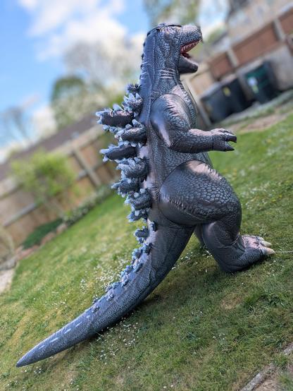 A grey inflatable Godzilla is seen from the side in a garden with an artificially blurred background. It has back skutes and a long tail.