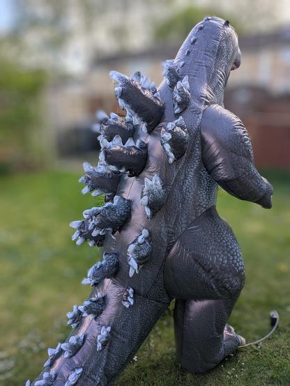 A grey inflatable Godzilla is seen from the back in a garden with an artificially blurred background. This view shows its back skutes/spikes.