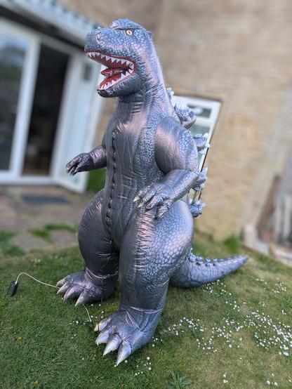 A grey inflatable Godzilla is seen from the front three-quarters view outside a house with an artificially blurred background.