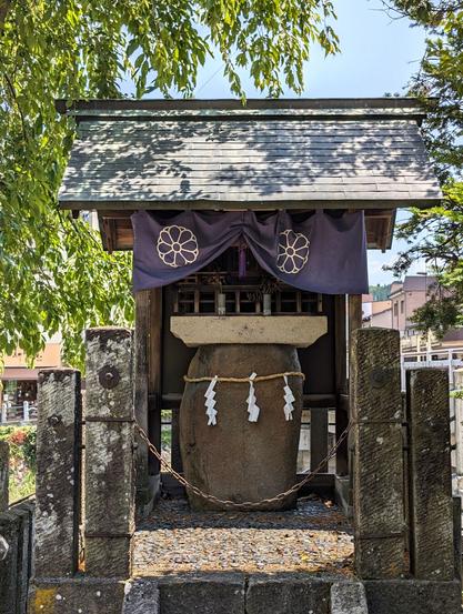 A rock enshrined in a little shrine. There are sacred ropes around it and purple curtains with a local emblem painted on them. It is the peak of the day in summer, with the hot sun rays hitting right from above.