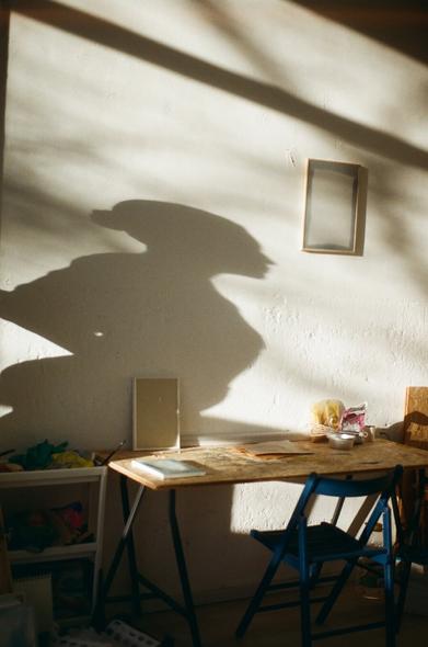 This is a photo, taken with an analog camera, taken inside an art studio. We see the desk of an artist and on the wall, leaning above the desk, the shadow of a real-size sculpture of a woman.