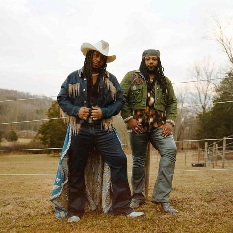 On the left, a Black man with long locks poses in a white cowboy hat and denim suit with golden fringe. He wears elaborately embroidered chaps. On the right, his twin wears a green denim cropped jacket, bell bottoms, shiny cowboy boots, and a red and black shirt. They stand on a rural ranch with a low hill in the background.