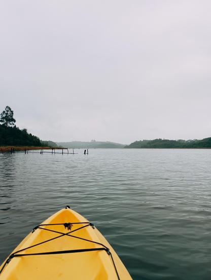 Vista desde la proa de un kayak amarillo navegando en un lago tranquilo bajo un cielo gris.  A lo lejos, se ven líneas de árboles oscuros en ambas orillas.  En el agua, cerca de la orilla izquierda, hay una estructura de madera sumergida, parcialmente visible. La atmósfera es serena y algo brumosa.