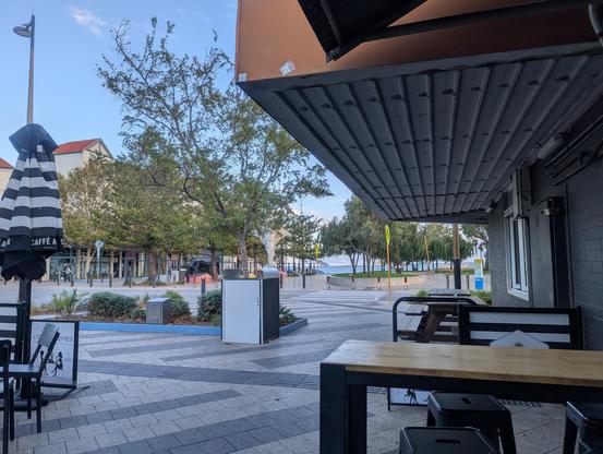 Outdoor view from a covered cafe seating area, looking towards a street with trees and benches. A large striped umbrella is visible. The area is paved, and buildings with red roofs are in the background. The sky is clear with a few clouds.