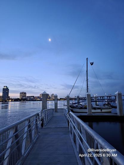 Early morning deep blue sky. Illuminated by a crescent moon above a boat dock gangway leading to a sailboat tied up on the river.