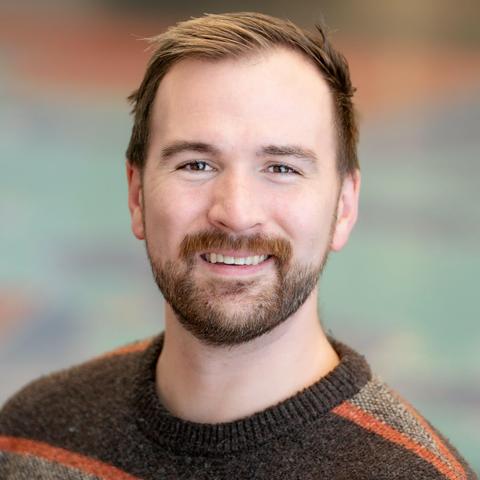 Color headshot photo of Eric Raglin, a fair-skinned man with short dark hair sun-bleached on the top and a neatly trimmed dark beard and mustache. He wears a brown-and-orange striped pullover sweater. He grins at the camera, brown eyes alight with…mischief? Devilishness? Something, though.