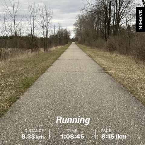 A concrete running path stretches into the distance, flanked by sparse trees and grass. Welcome to Michigan Rails to Trails in the early spring. The image displays running stats: a distance of 8.33 km, time of 1:08:45, and a pace of 8:15/km