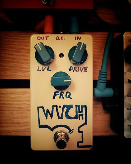 A guitar pedal with hand drawn labels and blue knobs sits on a wooden slat pedalboard