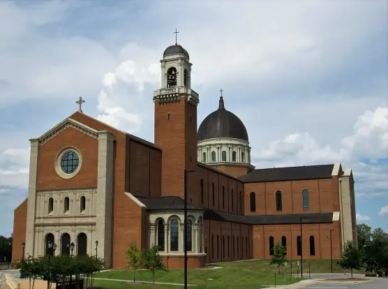 The image depicts a large brick building that appears to be a church or cathedral. It features a prominent dome, a bell tower with a clock, and several large windows. The structure has a mix of architectural styles, with elements that suggest it may be relatively modern. The surroundings include a green landscape and a cloudy sky.

Image Credits: Wikimedia / Farragutful / CC BY-SA 4.0
