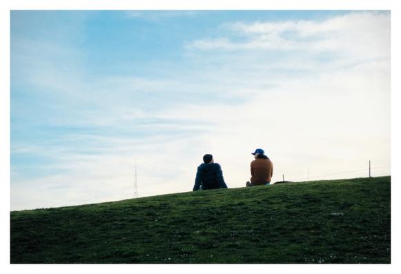 Two people sitting on grass, talking, with the sky above them.