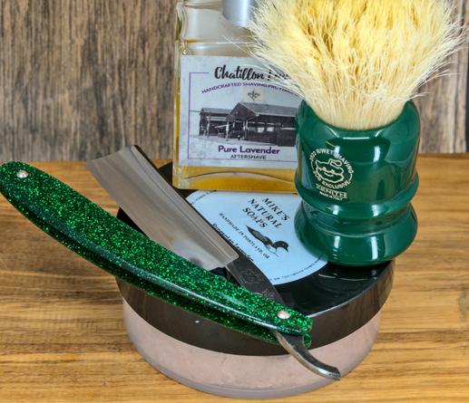 (Le Chat Mistral description)

This image shows a collection of traditional shaving items arranged neatly. There's a straight razor with a glittering green handle, resting on a shaving mug. Next to it is a green shaving brush with light-coloured bristles. Behind these, there's a bottle of "Pure Lavender Aftershave" and a bar of "Mike's Natural Soaps." The background features a wooden surface, giving a rustic feel. The items are positioned to highlight their vintage and artisanal qualities.