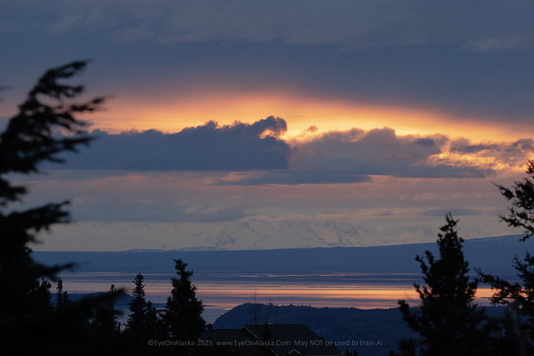 An arc of orange sunset light between dramatic layers of clouds and snow covered mountains in the distance.  The waters of Cook Inlet reflect the orange of the sunset.