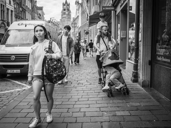 The image depicts a black and white street scene in Bruges, Belgium. In the foreground, a woman walks towards the camera, carrying a large patterned backpack. She is wearing a white hoodie, denim shorts, and white sneakers. To her right, a man is pushing a stroller with a child seated inside, while another child is perched on his shoulders. The background features a cobblestone street lined with buildings.