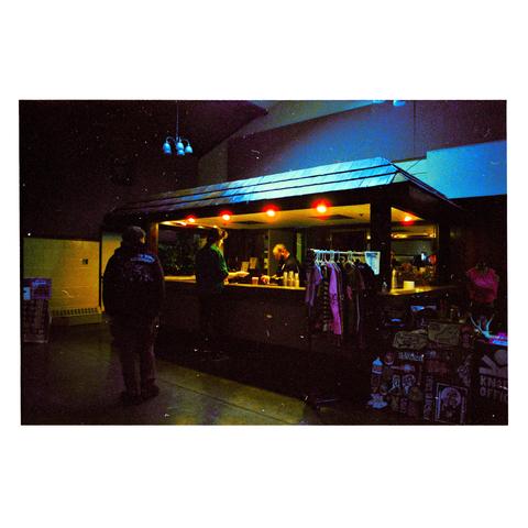 a film photo. in a dimly lit firehouse venue, a bar sits at the back of the main room. a man in a green shirt is at the counter, purchasing a drink from the bartender. someone stands behind the green shirt guy, looking on curiously. warm light emits from under the bar.