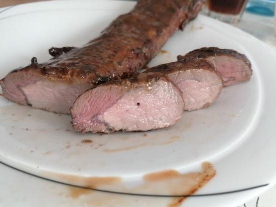 Das Bild zeigt ein geröstetes Stück Fleisch, wahrscheinlich Schweinebraten, auf einem weißen Teller. Das Fleisch ist in dünne Scheiben geschnitten, die die perfekte Röstung mit einem rosa Inneren und einer knusprigen, braunen Außenseite zeigen. Die Scheiben sind ordentlich angeordnet, und einige Fleischstücke sind noch ungeschnitten, was die Röstung und das gutes Zubereitungsergebnis deutlich macht. Auf dem Teller sind Fleischflüssigkeiten und Krümel zu sehen, was auf die Röstung hinweist. Im Hintergrund ist ein Glas mit einer dunklen Flüssigkeit, möglicherweise eine Sauce oder ein Getränk, zu erkennen. Der Teller hat einen schmalen schwarzen Rand, der das Fleisch hervorhebt.