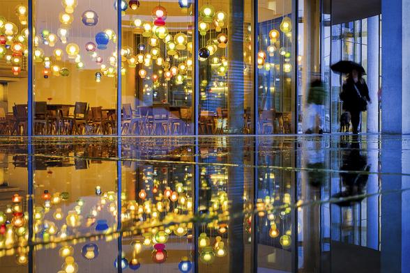 Symmetrical reflection shot in a puddle on the pavement of two people passing by a coffee storefront at blue hour in Berlin, on a rainy day. The coffee shop is bathed in golden light and decorated with multiple glass bubbles of different sizes and colors. The people are arriving from a corner of the image where the dark blue light evening light reflects on the windows of the next shop which light is switched off. One of the person is holding an open umbrella.
