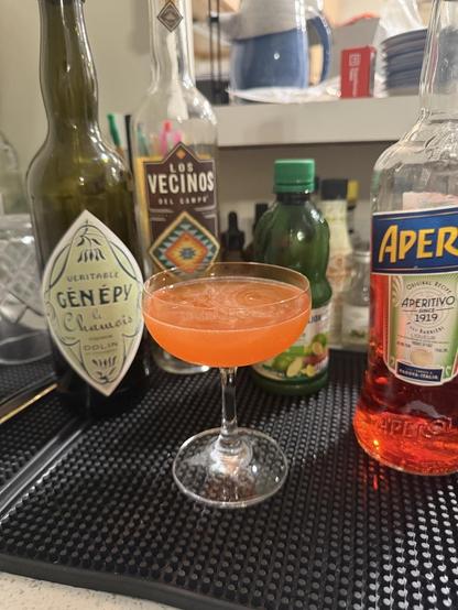 A light orange tinted drink in a coupe glass sits on a black bar mat. In the background, it is surrounded by the bottles of ingredients: Aperol, Mezcal, Génépy, and lime juice. I love an equal parts cocktail!