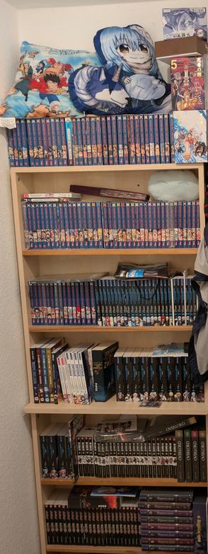 Das Bild zeigt eine Bücherregal-Einrichtung, die mit einer Vielzahl von Manga-Bänden gefüllt ist. Die Bücher sind in blauen Einbänden mit farbigen Illustrationen auf den Rücken geordnet, was auf eine Serie hinweist. Auf der obersten Regalbrett liegen zwei Kissen mit Anime-Motiven, einmal die Strohhut Piraten aus One Piece und einmal Rimuru aus Meine Wiedergeburt als Schleim in einer anderen Welt. Die Bücher sind in verschiedenen Reihen angeordnet, und einige sind in transparenten Schutzhüllen. Auf den Regalen sind auch einige andere Bücher und Gegenstände zu sehen, darunter eine Tasche und ein Buch mit dem Titel "Overlord". Die Bücherregale sind aus Holz und befinden sich an einer weißen Wand.