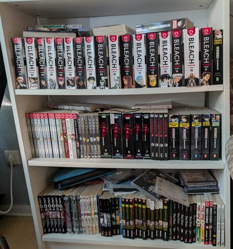 Das Bild zeigt ein Bücherregal mit mehreren Regalen, die mit Manga-Büchern und Comics gefüllt sind. Auf dem obersten Regal befinden sich mehrere Bände der Manga-Serie "BLEACH", die in Reih und Glied aufgereiht sind. Die Bücher haben schwarze und rote Einbände mit dem Titel "BLEACH" in großen weißen Buchstaben. Darunter sind weitere Manga-Bände in verschiedenen Farben und Designs zu sehen, darunter einige von "CLAMP" und "WANPANMAN". Das mittlere Regal enthält eine Mischung von Manga, insbesondere "xxxHolic", "Detektiv Conan Special Edition" und "Death Note". Das unterste Regal ist mit weiteren Manga-Büchern und einigen DVDs oder CD-ROMs gefüllt, darunter das Spiel "Age of Mythology" und verschiedene Bände des Manga "Dimension W". Einige Bücher sind unordentlich aufgestapelt. Das Regal ist weiß und steht an einer grauen Wand, an der ein Steckdosenanschluss zu sehen ist.