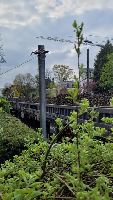 Hochkant in Farbe. Links im Bild eine niedrige, grüne Hecke, direkt rechts daneben die Gleise, auf denen ein, mit Gleisschotter beladener, Güterzug steht. Im Hintergrund mehrere Mehrfamilienhäuser, Laubbäume und ein Bagger. Der Himmel ist wolkig.