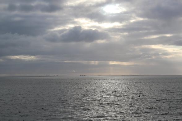 Landschaftsaufnahme der Nordsee. Untere Bildhälfte graues Meer mit Sonnenreflektion. Obere Bildhälfte abwechslungsreich und wolkenverhangen der graue Himmel mit Sonnendurchbrüchen. Am Horizont Warften einer Hallig. Im Wasser als kleines Detail eine Boje.