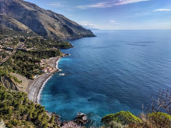 The image captures a scenic coastal view. A rugged mountain range dominates the left side of the frame, its slopes covered with sparse vegetation. The deep blue sea stretches out to the horizon, its surface reflecting the clear sky above. A small village nestles along the coastline at the base of the mountains, with buildings appearing as tiny red and white specks. A narrow, winding road hugs the shoreline, connecting the village to other areas. The foreground features a black sand beach, contrasting with the turquoise waters. The overall impression is one of natural beauty and tranquility.