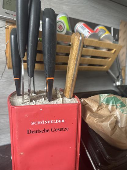 Eine Sammlung deutscher Gesetze als Messerblock hält mehrere Messer, darunter zwei mit Holzgriffen, auf einer Küchenarbeitsplatte.