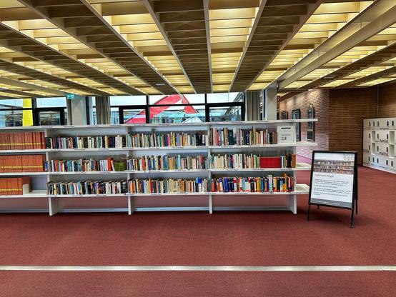 Ein helles Bibliotheksinterieur mit Bücherregalen, die mit verschiedenen Büchern gefüllt sind. Ein Schild in der Nähe bietet Informationen über einen "Buchmarkt". Der Raum hat ein modernes Design mit großen Fenstern und einem roten Teppichboden.
