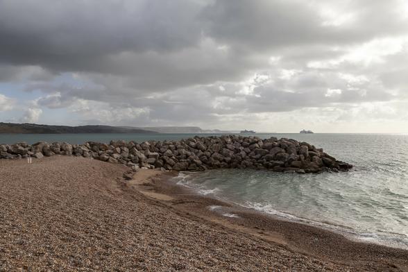 We're on a beach of fine, warm-toned pebbles, with the water's edge running from the bottom right of view to a small sand bank on which there are boulders amassed four or five deep above the surface to form a protective barrier a dozen metres long that stretches from left to right at around chest height. The sea is calm and grey-green in tone and it stretches to the horizon on the right but meets low, distant cliffs in hazy silhouette to the left. Far from shore, at sea there are three ships visible. One is an unladen cargo carrier and it's passing in front of one of the other two ships, dwarfed in size by them. They are both similar-looking cruise ships and it's just possible to make out a waved pattern of dark blue and white on their hulls with some lighter blue elements elsewhere, including the single funnel stack that each has. There are some patchy areas of blue sky in the clouds that are brightly lit by the sun to the right but carry the threat of rain in their dark masses to the left and above.