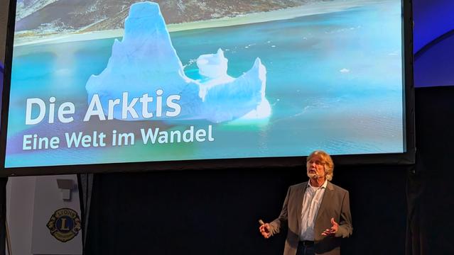 Arved Fuchs berichtet über die Arktis. Im Hintergrund ist eine Folie mit dem Text "Die Arktis. Eine Welt im Wandel" zu sehen.