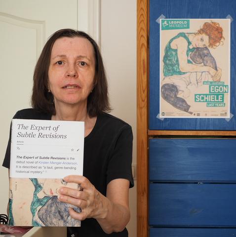 Eine Person hält ein Buch in die Kamera. Auf dem Buch-Cover ist ein Gemälde von Egon Schiele gedruckt. Auf dem Regal im Hintergrund klebt ein Plakat, welches dasselbe Gemälde abbildet.