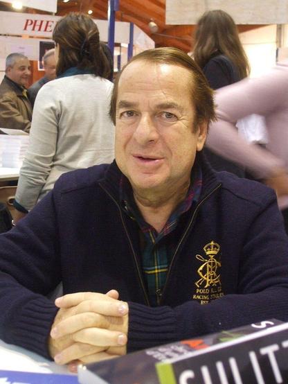 Paul-Loup Sulitzer, écrivain français, assis à une table lors de la foire du livre 2010 de Brive-la-Gaillarde. Il porte un pull bleu marine avec un logo doré sur la poitrine et une chemise à carreaux en dessous. Ses mains sont jointes devant lui sur la table. En arrière-plan, plusieurs personnes discutent près d'un stand de livres, ajoutant à l'ambiance animée de l'événement.