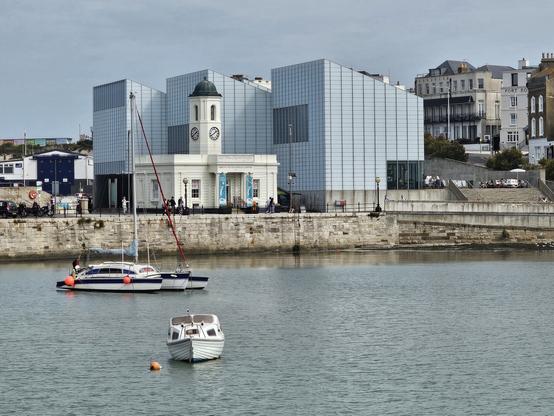 A view across water to a large, modern building on the quayside. It's basically three three white blocks with monopitch roofs. There are a couple of boats on the water, and an old harbourmaster's office next to the modern blocks.