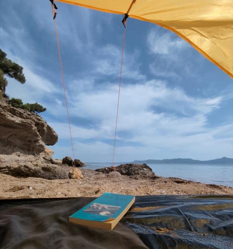 Photo taken from inside a yellow tent, showing a book, a beach, and the sea under a blue sky.